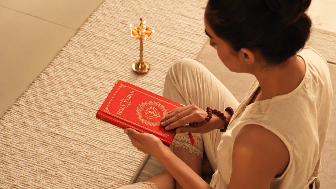 Žena sjedi na pletenoj prostirci i čita svete Vede uz mjedenu ayurvedsku uljanicu, za Art of Vedas